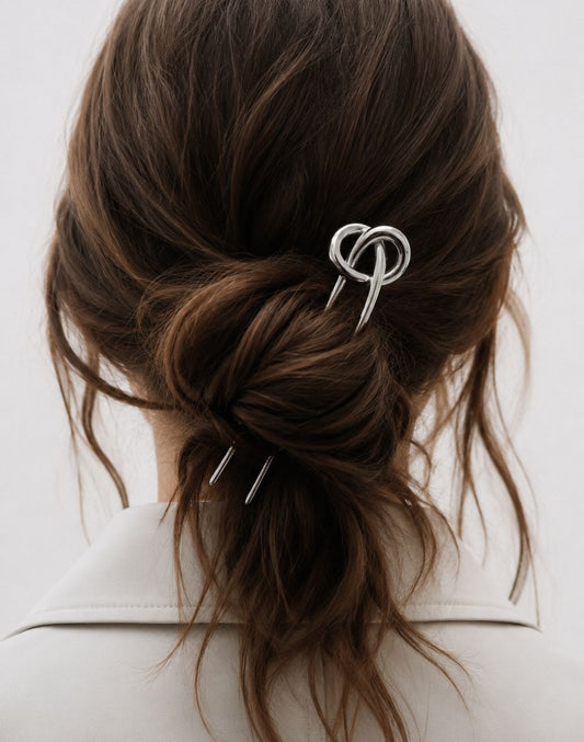 A silver metal hair claw clip with a knotted loop design, shown holding back a woman's brown hair in a low bun.