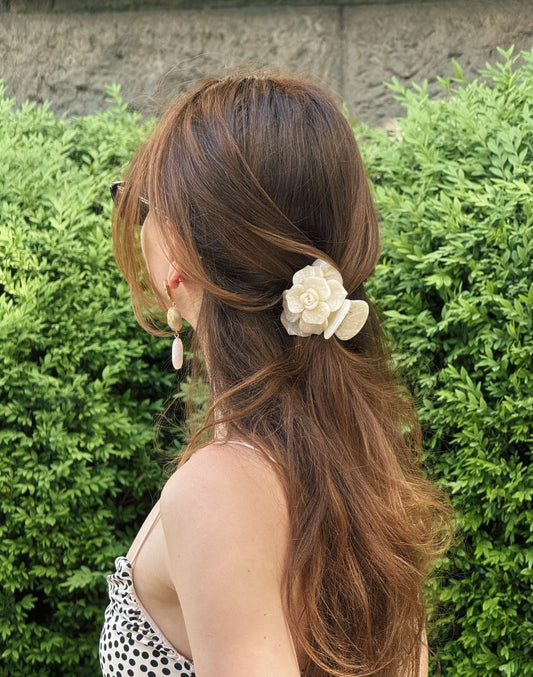 Woman with long brown hair tied back with a floral hair accessory, standing in front of green bushes.