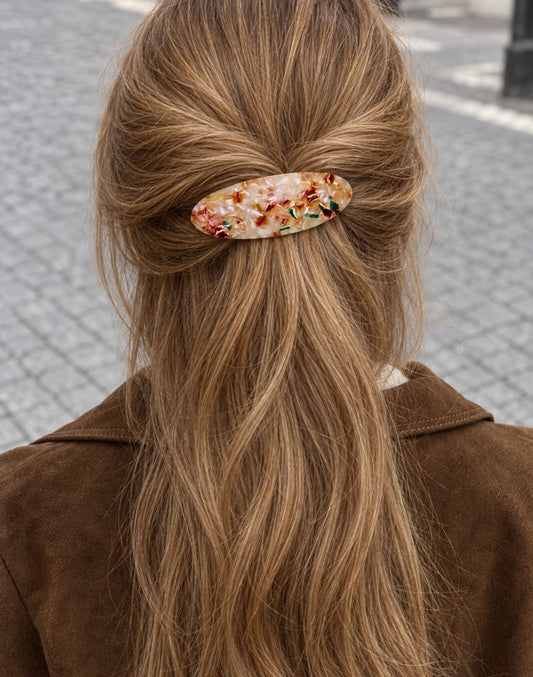A person viewed from behind with long light brown hair, wearing a brown jacket and holding a small oval hair clip with a multicolor floral pattern against the hair.