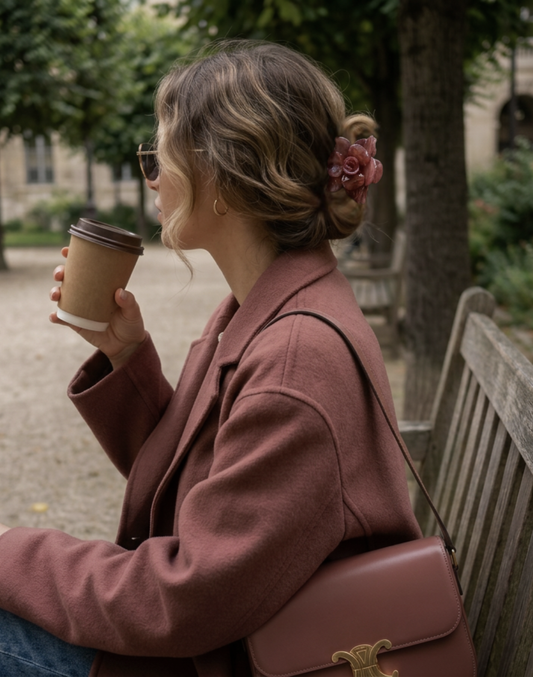 Woman in a pink coat sitting on a bench holding a coffee cup, with a blurred background of trees and a building.