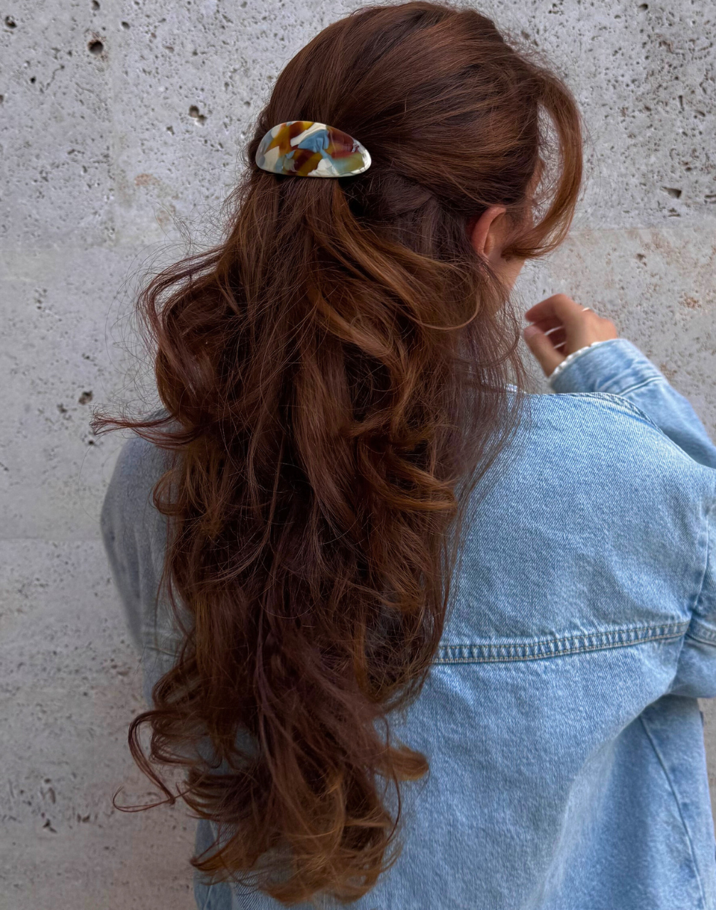 Person with long, wavy hair wearing a colorful hair clip against a concrete wall.