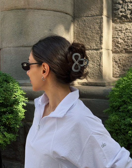 Woman wearing sunglasses and a white shirt with a floral hair accessory, standing against a stone wall.
