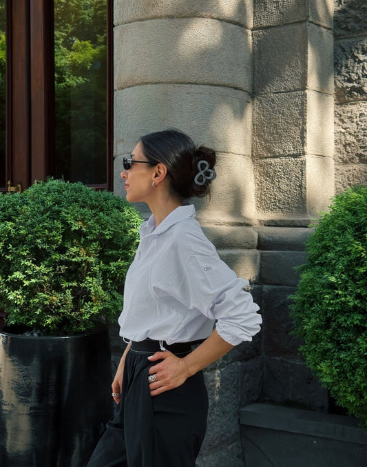 Woman in a white shirt and black pants standing against a stone wall with greenery.