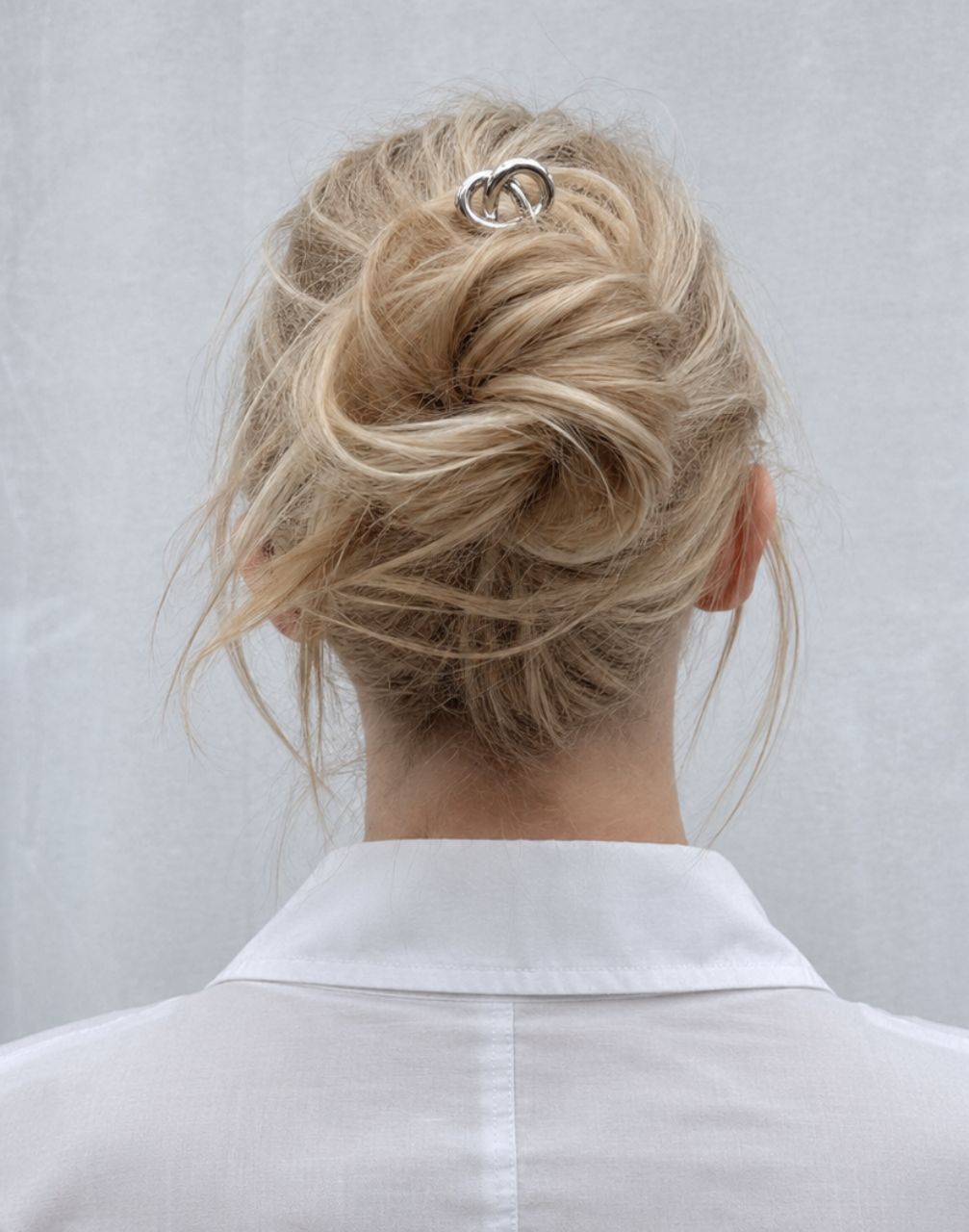A blonde woman viewed from behind wearing a white collared shirt and a silver interlocking rings hair clip in her messy bun.
