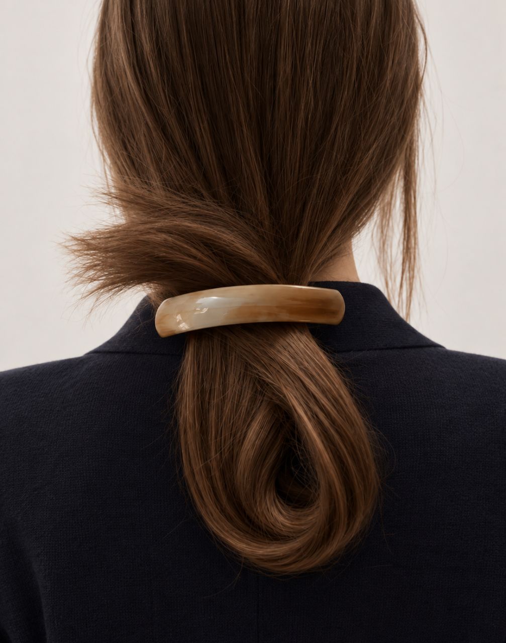 A close-up back view of a person with long brown hair tied in a low ponytail with a wide, curved tortoiseshell hair clip. The person is wearing a dark navy blazer against a plain light background.


