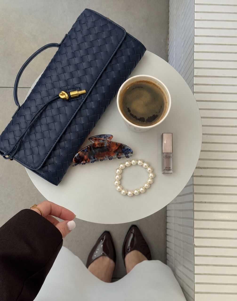 A navy blue woven leather clutch bag with a gold zipper pull, styled on a white side table alongside a coffee cup, a pearl bracelet, a tortoiseshell hair claw clip, and a lip product, with a person's hand and legs in brown pointed-toe flats visible at the bottom.