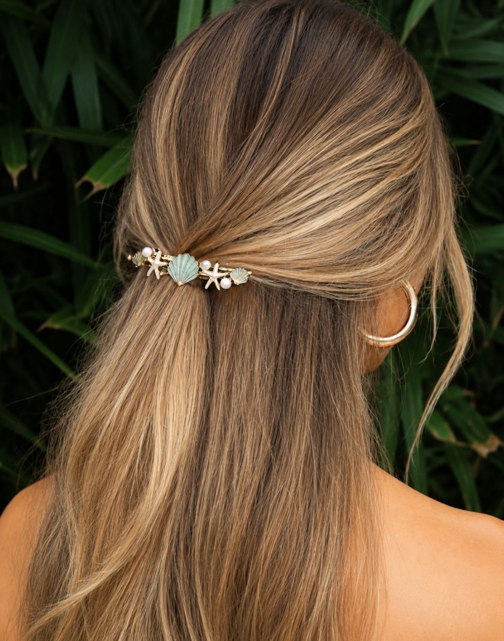 Woman with long, wavy hair wearing a decorative hair clip against a dark green leafy background