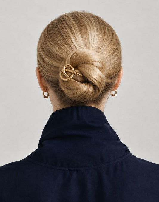 A blonde woman viewed from the back, wearing a navy blue high-neck top and a gold interlocking ring hair claw clip holding her hair in a low bun.